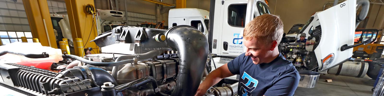 Student works on diesel engine with semi with open hood in background